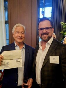 Two men in business attire smile at the camera indoors. The man on the left holds a piece of paper with notes and a glass of wine, while the man on the right wears glasses and a name tag that reads Ivan Ryan.