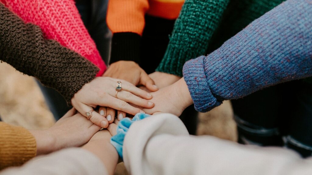 A group of people wearing colorful sweaters place their hands together in the center, showing unity and teamwork.
