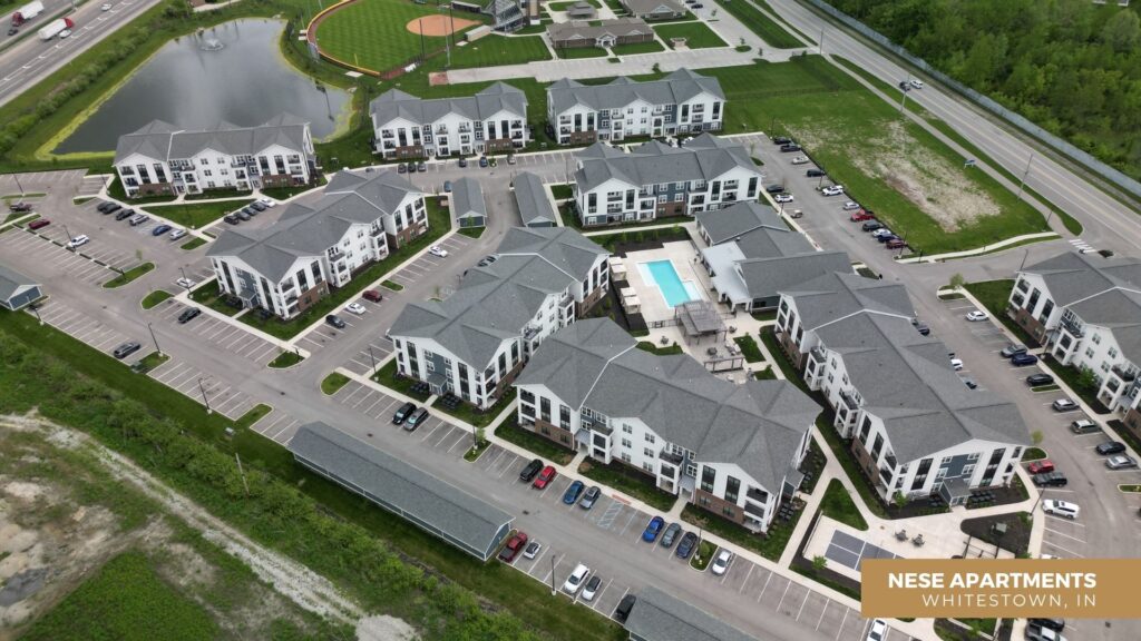 Aerial view of a modern apartment complex with multiple white buildings, a central swimming pool, parking lots, and nearby green spaces, including a baseball field and pond. Text overlay reads: NESE APARTMENTS, WHITESTOWN, IN.