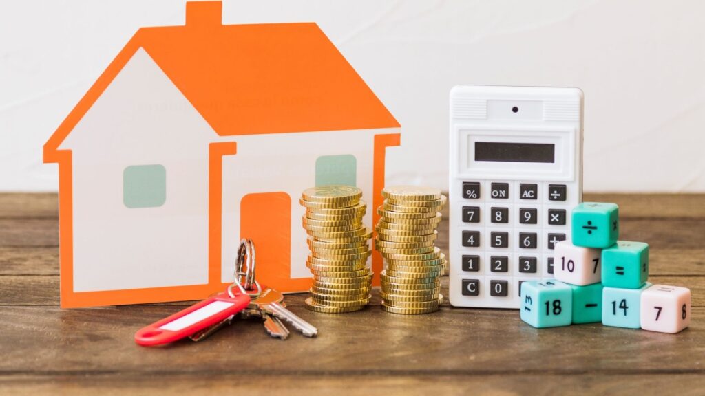 A paper house, stacked coins, a calculator, dice with numbers, and a set of keys with a red keychain are arranged on a wooden surface, symbolizing real estate, finance, and investments.