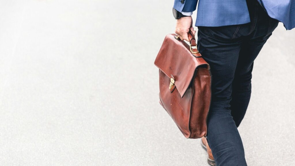 A person wearing dark pants and a blue blazer walks on a light-colored surface, carrying a brown leather briefcase by their side. Only the lower half of the person is visible.