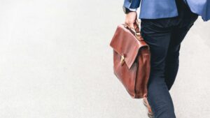A person wearing dark pants and a blue blazer walks on a light-colored surface, carrying a brown leather briefcase by their side. Only the lower half of the person is visible.