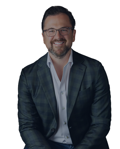 Smiling man with glasses and a beard, wearing a plaid dark blazer over a light dress shirt, sitting against a plain white background.