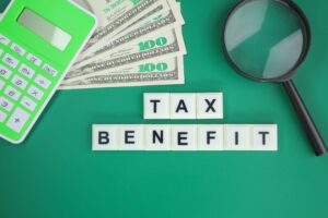 A green calculator, several 100-dollar bills, a magnifying glass, and letter blocks spelling TAX BENEFIT arranged on a green background.