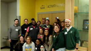 A group of people, some wearing festive holiday headbands and hats, pose and smile together in front of a yellow wall labeled Activity Center. A display case is visible on the right.