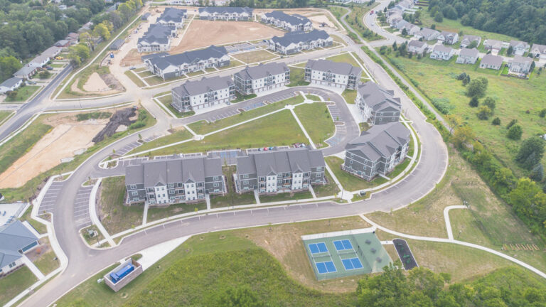 Aerial view of a suburban neighborhood with apartment buildings, curving roads, parking lots, green spaces, and tennis courts; surrounded by trees and undeveloped land.